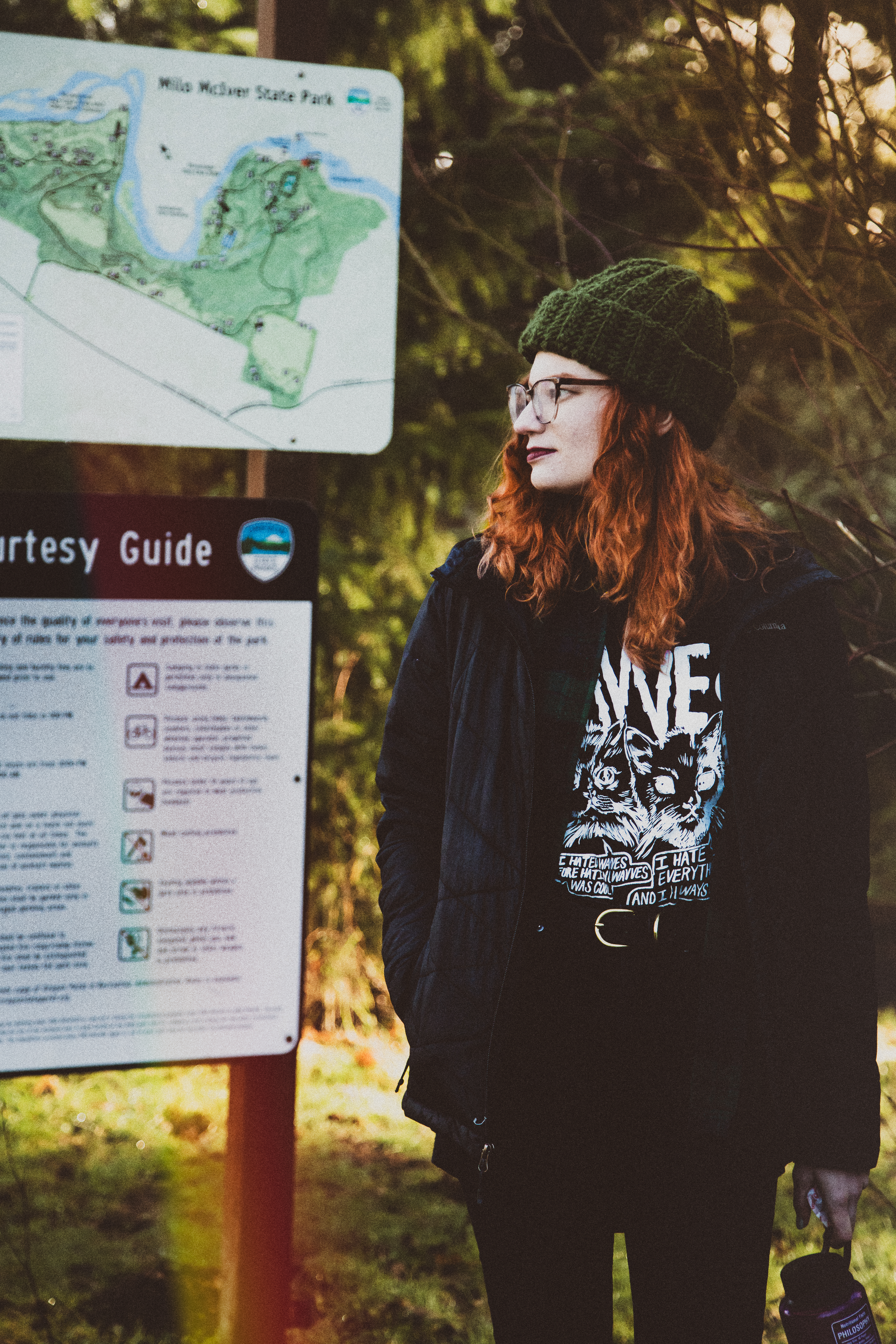 Kimberly Nachbur, a white woman with curly red hair, stands in a forest next to a trailhead sign. She is looking to the left, and she is wearing a green beanie, black rain jacket, black jeans, and a graphic t-shirt.